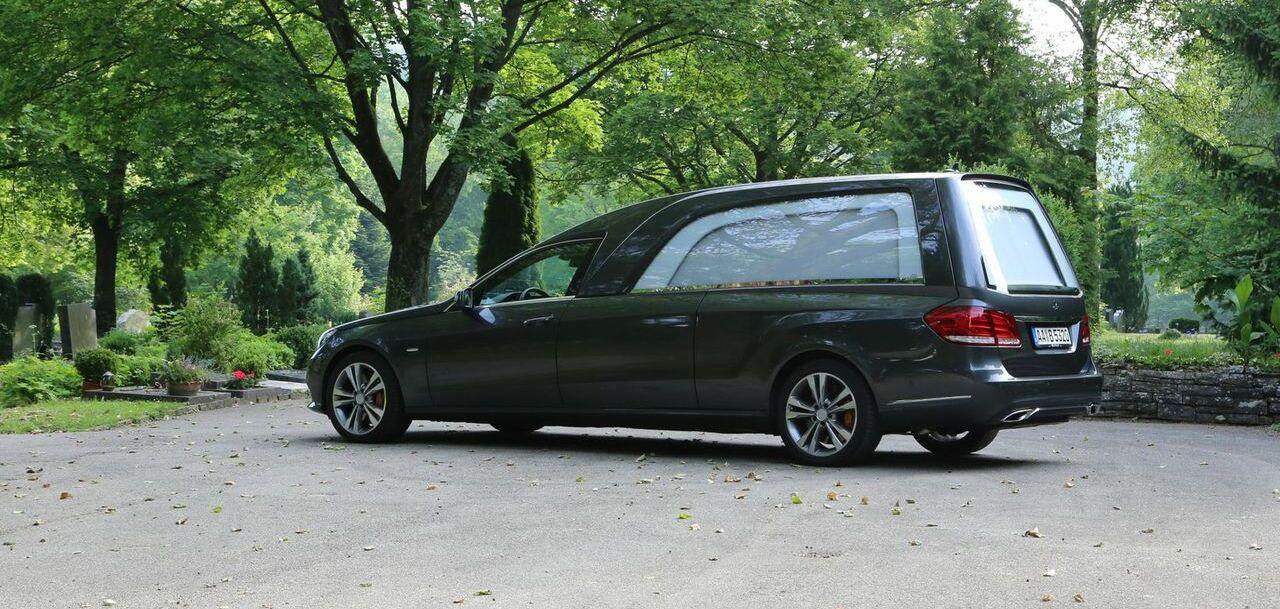 Dunkler Leichenwagen parkt auf einem Weg zwischen Bäumen auf einem Friedhof, neben einer Bank.