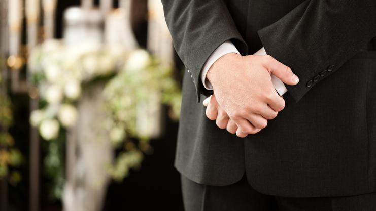 Ein Mann in einem Anzug steht mit verschränkten Händen vor einer dekorierten Kulisse mit Blumen und Kerzen.