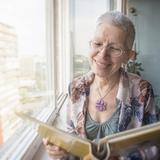 Lächelnde ältere Frau mit kurzen grauen Haaren schaut sich ein Fotoalbum an am Fenster mit Blick auf eine Stadtlandschaft.