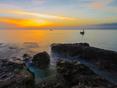 Sonnenuntergang über ruhigem Wasser, Felsen im Vordergrund und ein Boot entfernt auf dem Meer.